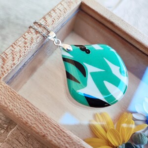 Puede incluir: Un collar con un colgante en forma de lágrima con un diseño abstracto verde, blanco y negro. El colgante está en una caja de madera con tapa de vidrio.