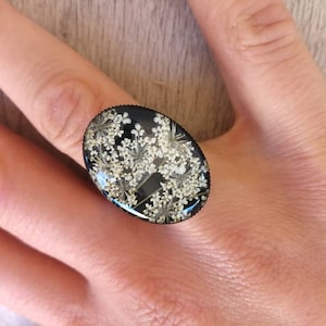 May include: A black ring with a clear oval-shaped resin top featuring a pressed white flower design.