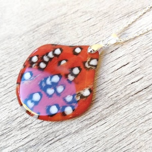 Puede incluir: Un collar con un colgante en forma de lágrima de resina roja y blanca. El colgante está unido a una cadena plateada.