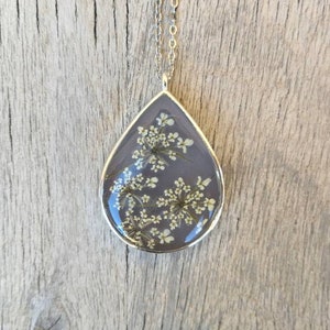 May include: Silver-toned necklace with a teardrop pendant. The pendant features a gray background with delicate white flowers encased in clear resin. The pendant is approximately 2 inches long and hangs from a delicate chain.