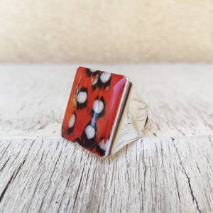 Könnte beinhalten: Ein silberner Ring mit einem quadratischen, rot, schwarz und weiß gemusterten Stein.