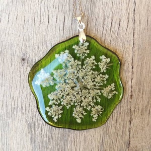 May include: A green resin pendant necklace with a white flower pressed inside. The pendant is shaped like a teardrop and has a silver chain.