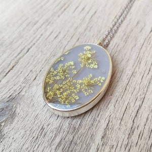 May include: A silver chain necklace with an oval pendant featuring pressed yellow flowers encased in clear resin. The pendant has a light gray background.