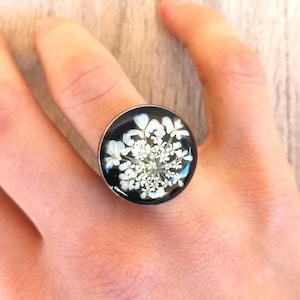 May include: A silver ring with a black background and a white flower pressed inside the resin.