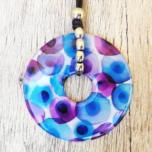 May include: A round pendant necklace with a white background and a pattern of blue and purple watercolor circles. The pendant is attached to a black cord with silver beads.