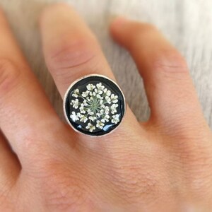 May include: A silver ring with a black background and a white flower pressed inside.