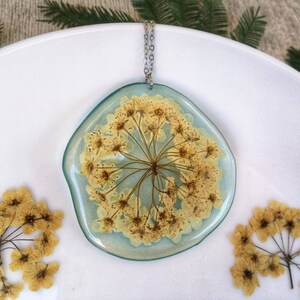 Large irregular pendant in blue transparent resin, with spirea flower inserted. H5,3xL5,3 cm approx. Stainless steel chain.
