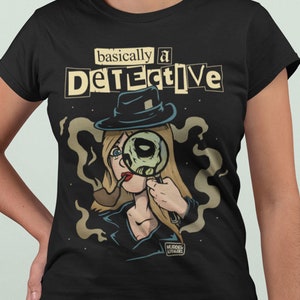 Peut inclure: T-shirt noir avec un graphique d'une femme portant un chapeau et tenant une loupe. Le texte "basically a DETECTIVE" est imprimé sur le t-shirt.