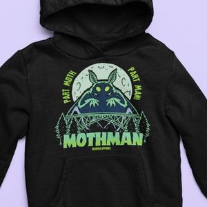 Op de afbeelding: Zwarte hoodie met een groene en witte afbeelding van een mot-achtig wezen met de tekst "Part Moth Part Man" en "Mothman" met de woorden "Murder Apparel" eronder.