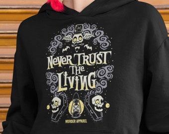 Puede incluir: Sudadera con capucha negra con un estampado gráfico de un personaje espeluznante con el texto "Never Trust The Living" y las palabras "Murder Apparel" en la parte inferior.