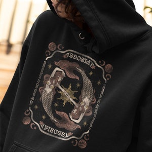 Puede incluir: Sudadera negra con un estampado gr&aacute;fico de dos sirenas con colas entrelazadas. El gr&aacute;fico est&aacute; rodeado de un c&iacute;rculo con las palabras "PISCES", "surrenders to the flow of life" y "a gifted guide for the wounded".