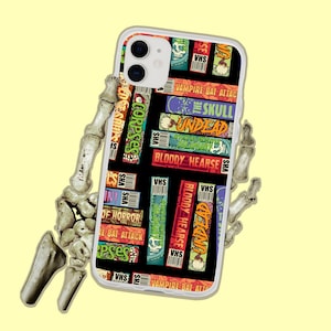 May include: A white phone case with a colorful design featuring various horror movie titles and VHS tapes. The titles include "Corpses", "The Skull", "Undead", "Bloody Hearse", and "Vampire Bat Attack".