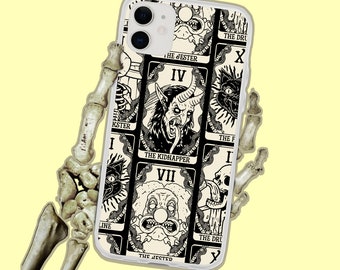 May include: A clear phone case featuring a tarot card design with black and white illustrations. The cards include images of a horned demon, a cat, and a clown. The phone is held by a skeletal hand against a yellow background.