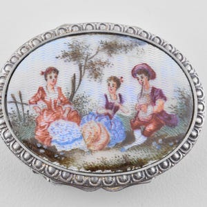 Op de afbeelding: Ovale zilveren sieradendoos met een geschilderde scène van drie figuren in klederdracht. De figuren bevinden zich in een landschap met een boom en een hek. De doos heeft een decoratieve zilveren rand.