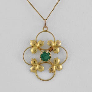 Op de afbeelding: Een goudkleurige hangerketting met een bloemmotief. De hanger heeft vier gestileerde bloemvormen rondom een centrale, ronde, groene edelsteen. De ketting is ook goudkleurig.