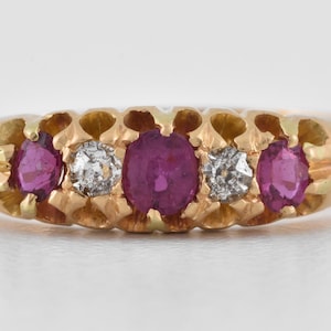 Może przedstawiać: Złoty pierścionek z pięcioma kamieniami szlachetnymi. Pierścionek ma dwa rubiny o owalnym szlifie, dwa małe diamenty i centralny owalny rubin. Oprawa ma dekoracyjny, ażurowy wzór.