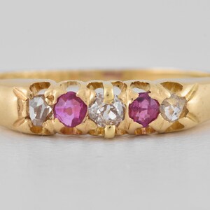 May include: A gold ring featuring a row of five stones set in a metal band. The setting includes two clear, faceted stones on the outer edges, two vibrant pink stones positioned inward, and a single clear, faceted stone at the center.