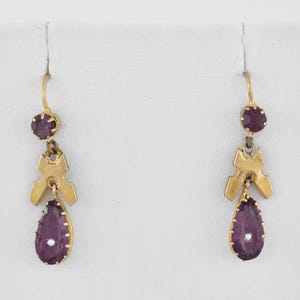 May include: A pair of gold drop earrings featuring a small round purple gemstone at the top, a central gold geometric component, and a larger pear-shaped purple gemstone at the bottom with a tiny white accent bead in the center of each drop.