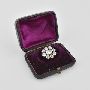 May include: A vintage brooch with a floral design, set in a black leatherette box with a purple velvet interior. The brooch features multiple clear gemstones arranged in a circular pattern, creating a sparkling effect.