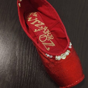 May include: Red sparkly pointe shoe with white and gold embellishments. The shoe is decorated with pearls and rhinestones. The inside of the shoe is lined with red fabric and has the words "ARD 70" written in gold.