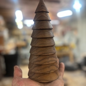 May include: A handcrafted wooden Christmas tree with a conical top and tiered, rounded layers. The tree is made of a dark brown wood, showcasing the natural grain. The tree is held in a hand, with a blurred workshop background.