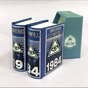 May include: Two miniature blue books with the title "1984" by George Orwell. Each book features a pyramid with an eye symbol. A matching teal box with the same symbol is also present.
