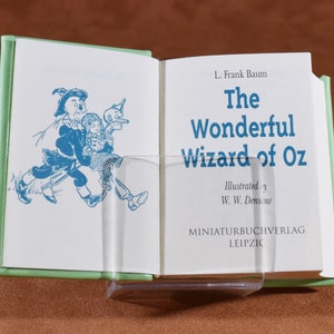 May include: An open miniature book with a light green cover, titled "The Wonderful Wizard of Oz" by L. Frank Baum. The book's pages display blue illustrations and text. The book is held open by a clear plastic stand.