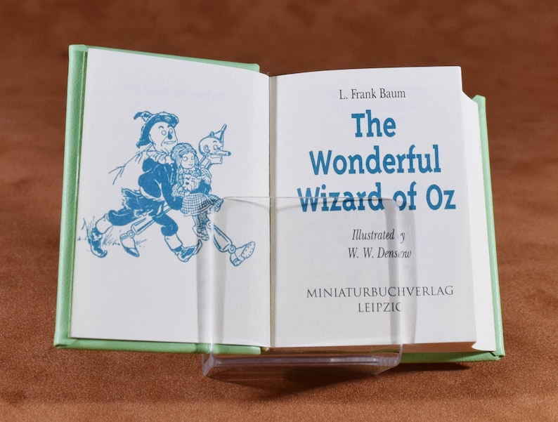 May include: An open miniature book with a light green cover, titled "The Wonderful Wizard of Oz" by L. Frank Baum. The book's pages display blue illustrations and text. The book is held open by a clear plastic stand.