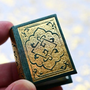 Peut inclure: Un petit livre avec une couverture verte et des détails dorés en relief. La couverture présente des motifs dorés ornés et des caractères arabes. Le livre est tenu dans une main, soulignant sa petite taille et son design complexe.