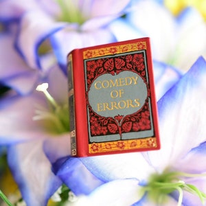 May include: A small red book with gold lettering on the cover. The title of the book is "Comedy of Errors". The book is sitting on a bed of white and blue flowers.