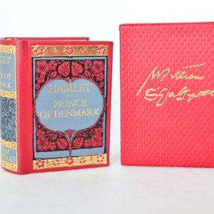 May include: A red leather-bound book with gold lettering titled "Hamlet, Prince of Denmark". The book is closed and sitting on a white surface. A red box with a gold inscription of "William Shakespeare" is next to the book.
