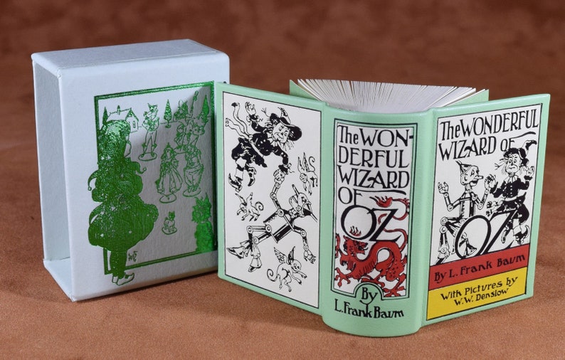 May include: A miniature book set of "The Wonderful Wizard of Oz" by L. Frank Baum. The book has a light green cover with illustrations and text. The set includes a matching box with a green illustration of characters.
