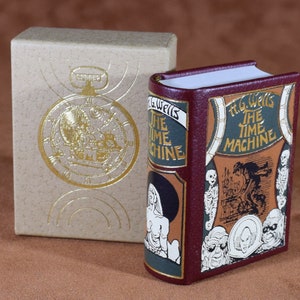 May include: A miniature book titled "The Time Machine" by H.G. Wells, with a burgundy cover and gold lettering. The book is in a matching box with a gold embossed pocket watch design. The book's cover features illustrations of characters and scenes.
