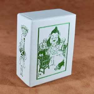 May include: A white box with green illustrations. One side features a drawing of a character holding a torch, while the other side depicts a scene with a dog-like character at a table with other figures. The box is on a brown surface.