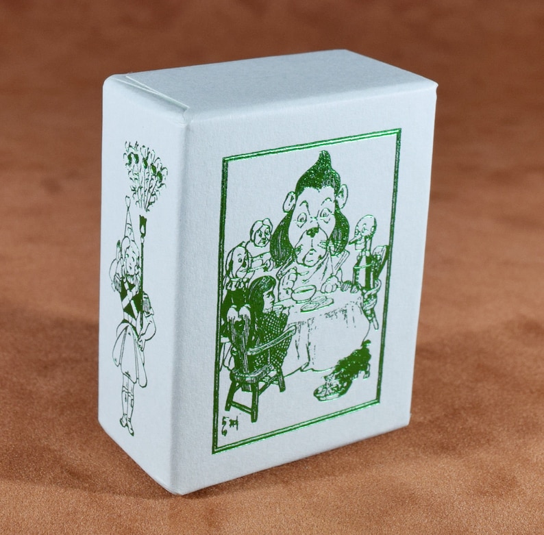 May include: A white box with green illustrations. One side features a drawing of a character holding a torch, while the other side depicts a scene with a dog-like character at a table with other figures. The box is on a brown surface.
