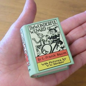 May include: A miniature book titled "The Wonderful Wizard of Oz" by L. Frank Baum. The book has a light green cover with illustrations of characters and the title in black. The book is held in a hand.