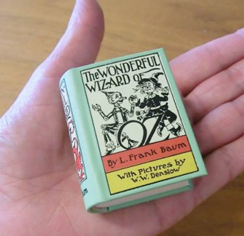May include: A miniature book titled "The Wonderful Wizard of Oz" by L. Frank Baum. The book has a light green cover with illustrations of characters and the title in black. The book is held in a hand.