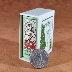May include: A miniature book titled "The Wonderful Wizard of Oz" in a white box, next to a U.S. quarter. The book has a red dragon illustration and the author's name, L. Frank Baum. The box has green illustrations.