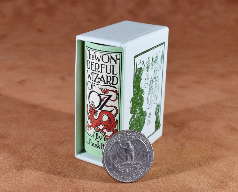 May include: A miniature book titled "The Wonderful Wizard of Oz" in a white box, next to a U.S. quarter. The book has a red dragon illustration and the author's name, L. Frank Baum. The box has green illustrations.