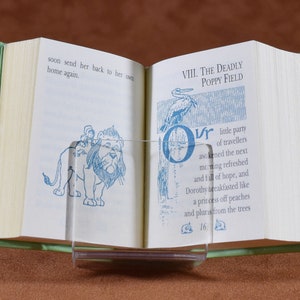 May include: Open book with green covers, held open by a clear acrylic stand. The pages display blue illustrations and text, including "VIII. THE DEADLY POPPY FIELD" and a drawing of a lion. The book is open to page 16.