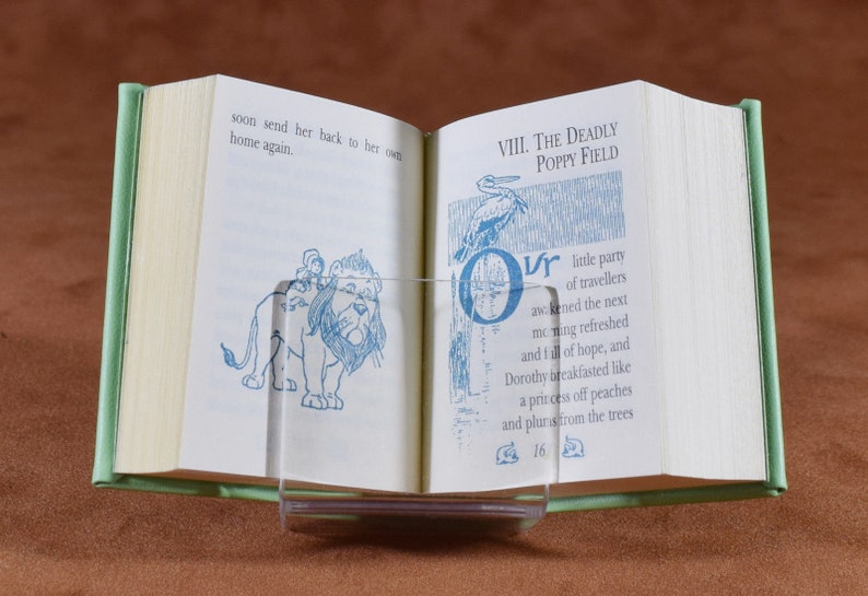 May include: Open book with green covers, held open by a clear acrylic stand. The pages display blue illustrations and text, including "VIII. THE DEADLY POPPY FIELD" and a drawing of a lion. The book is open to page 16.