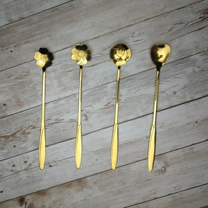 May include: Four gold-coloured metal stirring spoons with different decorative designs on the ends. The spoons are arranged in a row on a light wood surface.