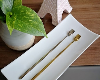 May include: Two long, thin spoons with rounded ends. One spoon is silver and the other is gold. The spoons are on a white rectangular plate.