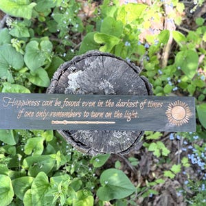 May include: A dark green wooden sign with the quote "Happiness can be found even in the darkest of times if one only remembers to turn on the light." The sign has a sun and wand design. The sign is resting on a tree stump surrounded by greenery.