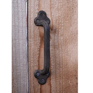 May include: Black metal door handle with a rounded rectangular shape and a decorative top. The handle is attached to a wooden door with screws.