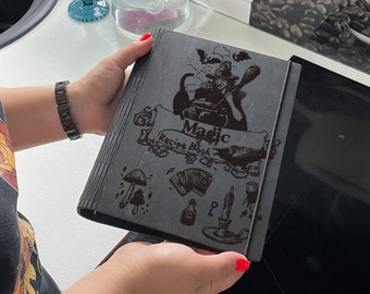 Può includere: Un libro di ricette in legno grigio scuro con le parole "Magic" e "Recipe Book" incise sulla copertina. La copertina presenta illustrazioni di una strega, funghi e altri elementi magici. Il libro è tenuto in mano.