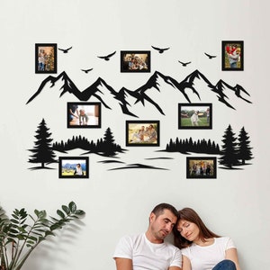 May include: A black silhouette of mountains, trees, and birds with 10 empty picture frames. The frames are arranged in a decorative pattern on a white wall. A couple is sitting on the floor in front of the wall.