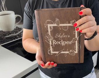 May include: A brown wooden recipe book with a black elastic band closure. The front cover is engraved with the text "Skylar's Recipes" and a design of kitchen utensils and food.