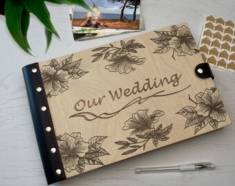 May include: A wooden photo album with a black leather cover and a floral design. The album has the words "Our Wedding" engraved on the front.