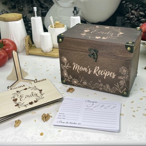 May include: A wooden recipe box with a floral design and the text "Mom's Recipes" and "Emily" engraved on the lid. The box is open and there are recipe cards inside. There is also a wooden recipe card holder with the text "Emily" engraved on it.